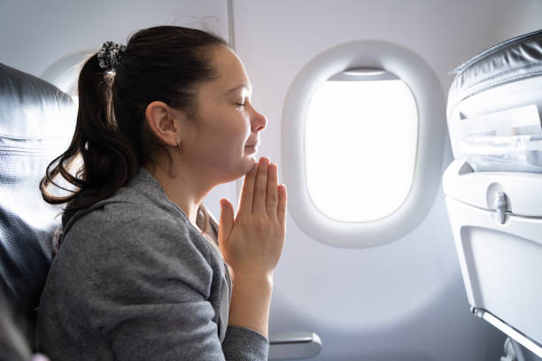 A woman sitting on an airplane seat, eyes closed, hands clasped in prayer, with sunlight streaming through the window.